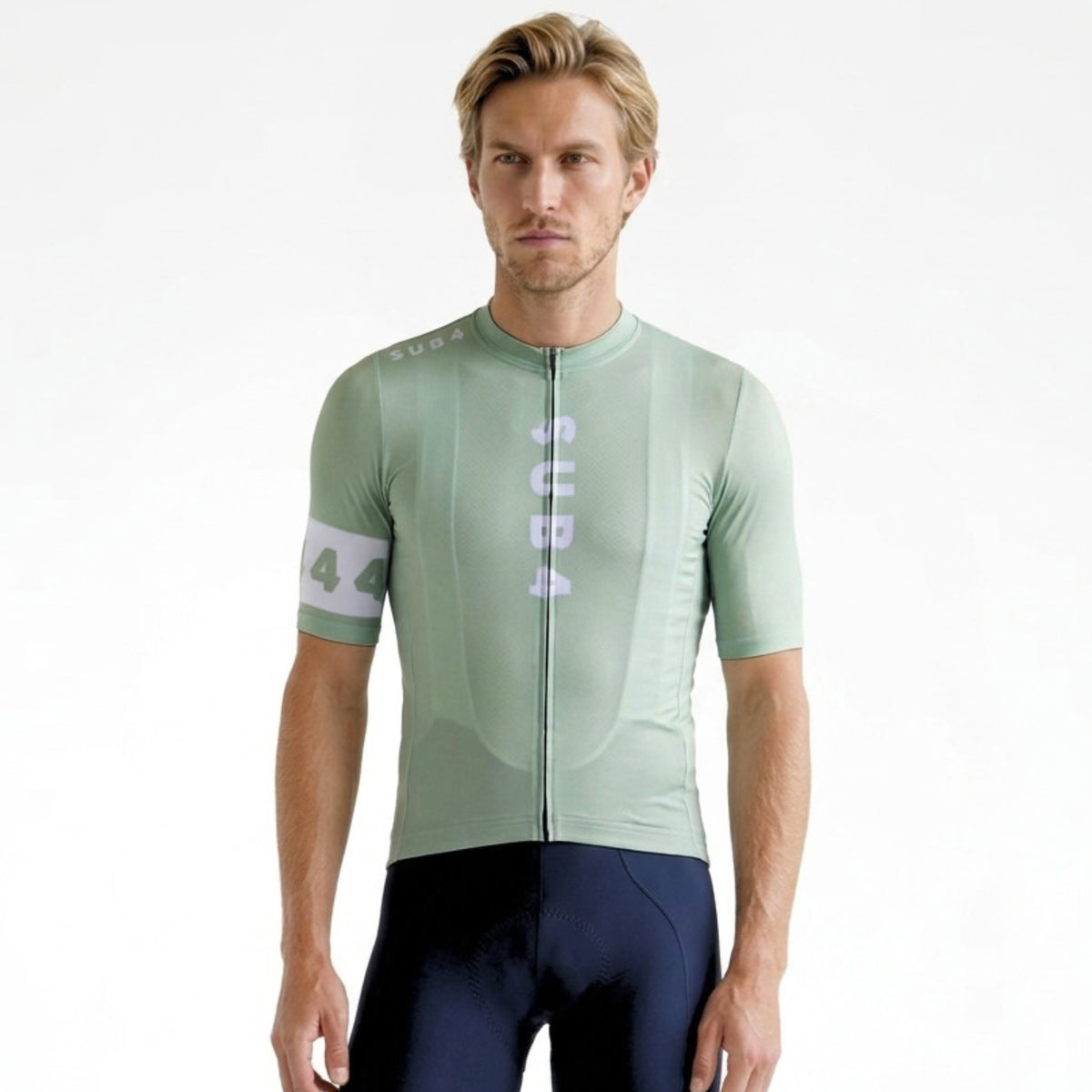 A male model wearing a light sage green short-sleeve cycling jersey with a front zipper and vertical 'SUB4' branding, paired with dark navy cycling shorts, against a plain white background.